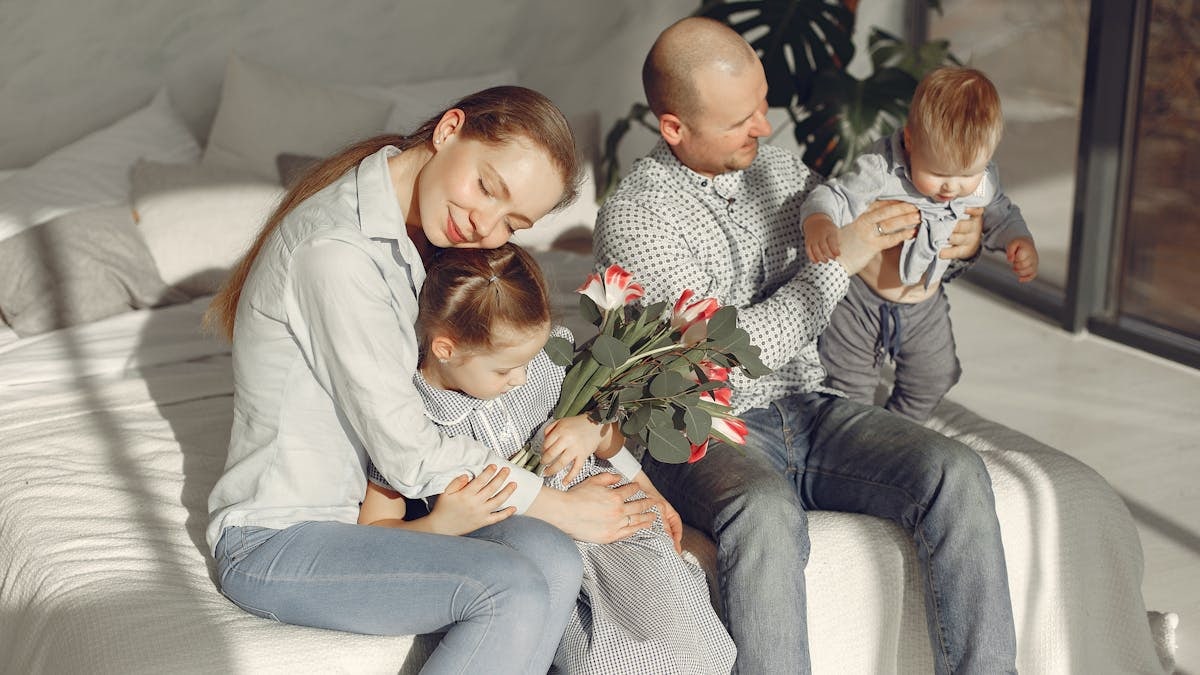 Cheerful parents with children wearing casual clothes smiling while playing with daughter and son in bedroom together during weekend at home